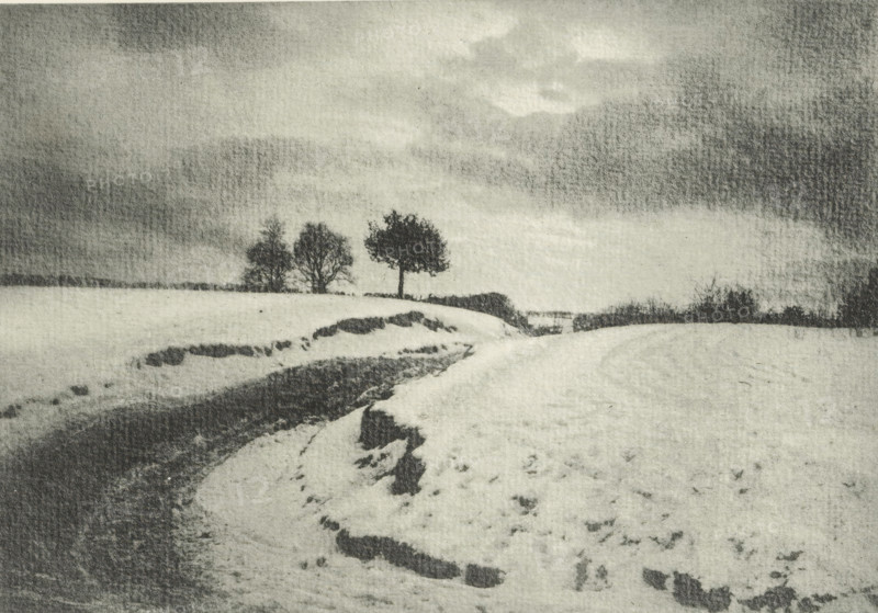 Landscape with country road in winter, Une route de campaign en hiver, photomechanical print, photographer: Charles Job, cliché maker: Fillon & Heuse, printer (person): Charles Wittmann, Paris, c. 1891 - in or before 1896, printer (person), paper, photoaquatint, height 133 mm x width 191 mm, road, path, low hill country, winter landscape, landscape symbolizing winter (the four seasons of the year), A grainy black and white landscape showing a snow-covered field with a curving snow-lined path or ditch leading toward the middle distance, dark patches of exposed ground along its edges, and a small group of bare trees on the horizon beneath a wide, cloudy sky.