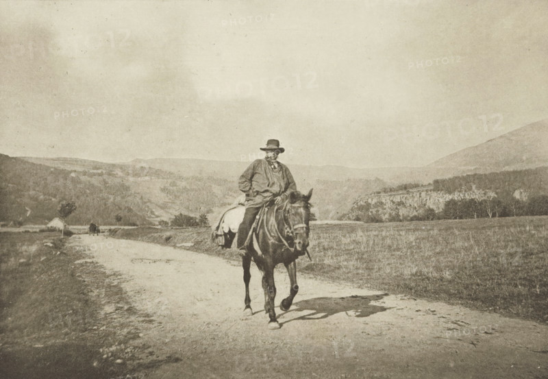 Retour du Moulin (Mont-Dore), Rider on a country road, Mont-Dore, photomechanical print, photographer: G. de Magnitot, cliché maker: Fillon & Heuse, printer (person): Charles Wittmann, Paris, c. 1891 - in or before 1896, printer (person), paper, photoaquatint, height 129 mm x width 185 mm, riding a horse, ass, or mule, rider, horseman, (high) hill, A sepia-toned scene shows a person wearing a wide-brimmed hat and loose outer garment seated on a saddled horse that is walking along a dirt road, with the rider holding the reins and looking slightly to the side; the road stretches into a pastoral landscape with grassy fields and low hills, scattered trees, and a small building near the roadway in the distance, while further hills and a cliff-like formation rise beyond and the sky above is pale and expansive.
