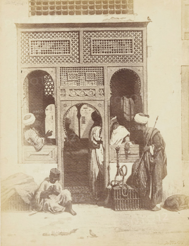 Photo reproduction of the painting 'A barber's shop in Cairo' by Willem de Famars Testas, photograph, reproduction, page, photographer: Binger & Chits, after painting by Willem de Famars Testas, c. 1866 - in or before 1871, Binger & Chits, Willem de Famars Testas, photographer, after painting by, paper, albumen print, height 190 mm x width 147 mm, barber, barbershop, A monochrome scene shows an ornate doorway with carved and latticed panels and arched openings; patterned screens fill the upper portion and decorative motifs frame the central archway. Several figures in flowing robes and head coverings stand and sit around the entrance, one seated on the ground with folded legs, another leaning near a window opening, and others clustered in and near the doorway. A tall figure holds a long staff and a coiled hose or pipe rests on a wicker crate beside them. Rounded sacks or bundles lie near the walls and a small bird is visible on the ground in front of the entrance.