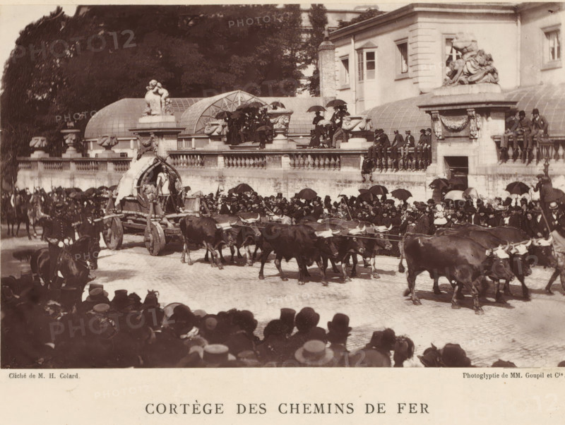 Nervian ox cart, at a parade for the railways in Brussels, Cortège des chemins de fer, chariot Nervien (Bruxelles 1885), page, photomechanical print, photographer: Hector Colard, Brussels, cliché maker: Goupil & Cie., Paris, 1885, printer (person), paper, Woodburytype, height 103 mm x width 161 mm, Print on unnumbered page., public festivities, four-wheeled vehicle drawn by more than three animals, festivities, corporative organization, guild, A wide street scene shows a procession with a team of oxen pulling a wooden wagon that has a curved canopy and figures standing on and around it, a large crowd lines the route with many people holding umbrellas and wearing hats, a stone balustrade and terrace above the street is occupied by seated and standing spectators, a building with classical ornamentation and sculptural figures sits behind the terrace, decorative stone urns and carved details adorn the balustrade, several mounted riders are visible among the crowd, and trees rise behind the...