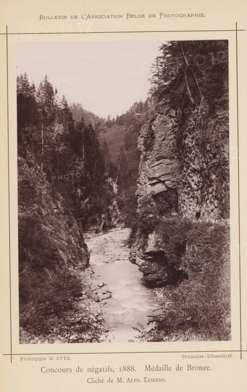 Rock formations in the Tamina valley, Switzerland, page, photomechanical print, inscription: 'Concours de négatifs, 1888. Médaille de Bronze.', photographer: Alphonse Leirens, Tamina Gorge, cliché maker: Wilhelm Otto, Brussels, 1886-09-07, printer (person), Taminakloof, pasteboard, paper, collotype, photoprint, height 170 mm x width 120 mm, Print on unnumbered page., brook, rock-formations, Tamina, A narrow river flowing between steep rocky cliffs and wooded slopes, with trees lining the ridges and growing on the cliff faces, a rocky overhang on the right side, vegetation and shrubs along the riverbanks, a distant hillside with scattered trees, and the photograph printed on a bordered page with a printed heading at the top and printed captions along the bottom.