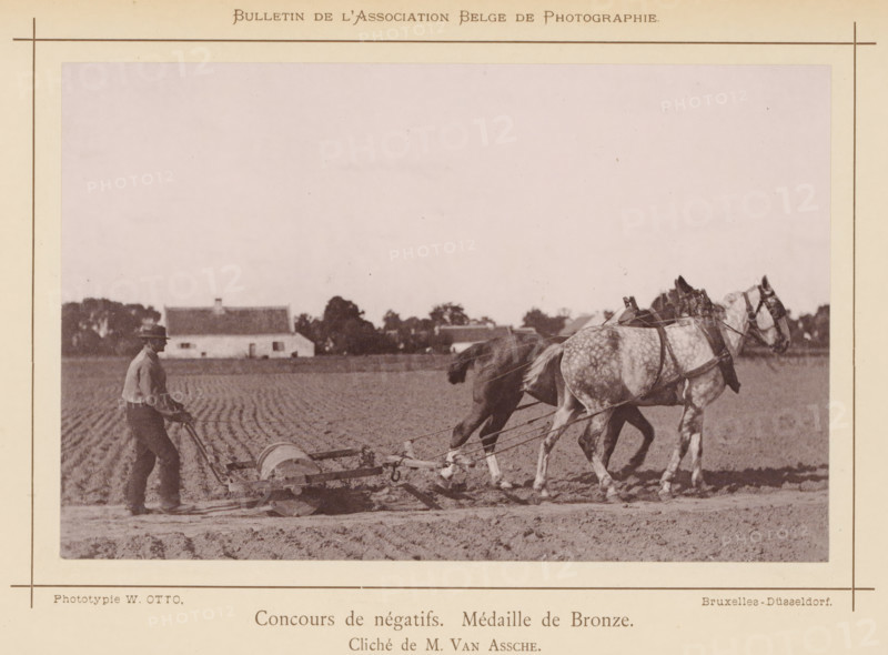 Farmer rolling his field, Le paysan au labour, page, photomechanical print, inscription: 'Concours de négatifs. Médaille de Bronze.', photographer: Guillaume van Assche, cliché maker: Wilhelm Otto, Brussels, c. 1883 - in or before 1888, printer (person), pasteboard, paper, collotype, photoprint, height 109 mm x width 169 mm, Print on unnumbered page., rolling, soil cultivation, A sepia-toned photograph shows a field scene with a man walking behind a plow while holding its handles, two horses harnessed side by side in front of the plow, and furrowed soil underfoot; low buildings and scattered trees appear along the horizon in the background, and the image is framed by a pale border with thin lines near the edges.