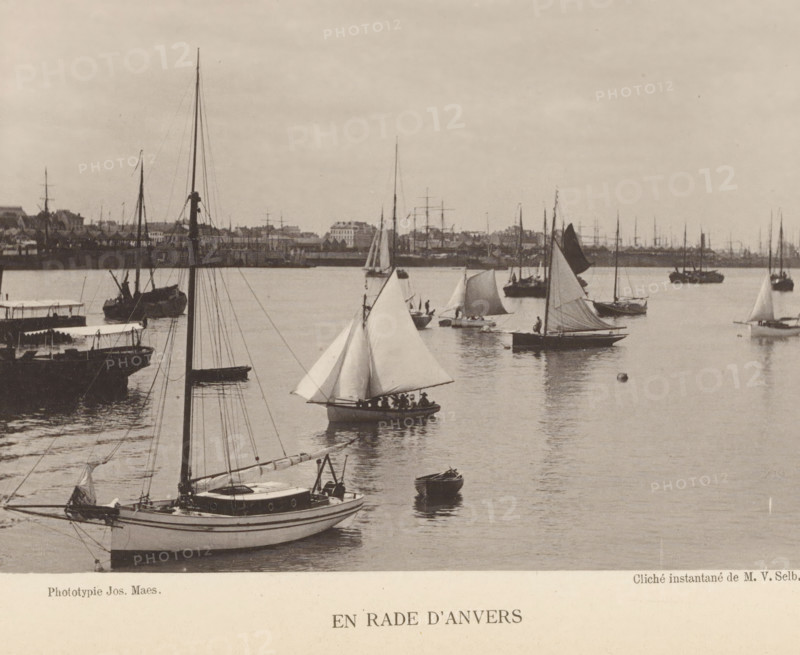 Sailing ships at the port of Antwerp, En rade d'Anvers, page, photomechanical print, inscription: 'Section d'Anvers - Exposition de 1889', photographer: Victor Selb, Antwerp, cliché maker: Joseph Maes, c. 1883 - in or before 1888, printer (person), paper, collotype, photoprint, height 115 mm x width 156 mm, Print on unnumbered page., harbour, sailing-ship, sailing-boat, A waterfront scene showing multiple sailboats and small boats on calm water with visible masts, rigging, and unfurled sails; a larger sailboat in the foreground with a cabin and visible deck fittings, smaller sailboats with triangular sails scattered across the water, and a pair of rowboats; reflections of the boats on the water surface; a distant shoreline with buildings and additional masts lining the horizon; a mostly overcast sky above.