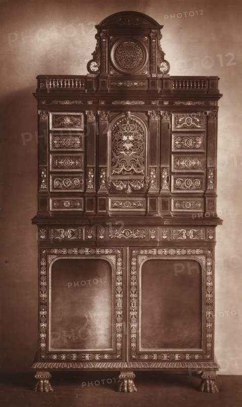Cabinet in the style of Louis XIII, from the collection of Christopher Beckett Denison, page, photomechanical print, number: '1868', c. 1880 - in or before 1885, paper, Woodburytype, height 195 mm x width 121 mm, Print on third unnumbered page after page 178., ornamental cupboard (wood and other phytogenic material), A tall ornate wooden cabinet photographed against a plain backdrop, featuring multiple stacked sections with carved moldings and decorative inlays; the uppermost portion has a rounded pediment with a circular carved medallion flanked by scroll-like ornaments and small balustrades, below which are rectangular panels with intricate scrollwork and floral motifs; the central section displays a recessed arched panel filled with elaborate symmetrical foliage and foliate patterns surrounded by fluted pilasters and framed drawers and panels bearing carved ornament; the lower section includes two large recessed door openings bordered by patterned inlay and narrow vertical columns o...