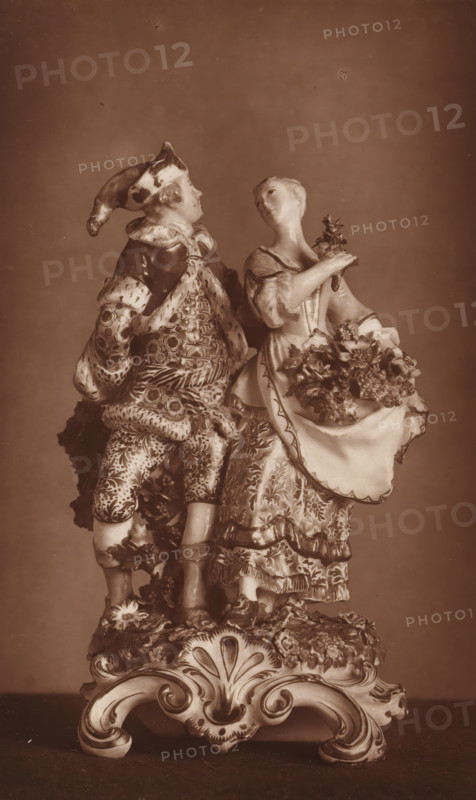 Two porcelain statues, depicting summer and winter, page, photomechanical print, number: '3101', after sculpture by anonymous, c. 1880 - in or before 1885, after sculpture by, paper, Woodburytype, height 194 mm x width 121 mm, summer, 'Aestas', 'Estate' (Ripa), winter, 'Hyems', 'Inverno' (Ripa), piece of sculpture, reproduction of a piece of sculpture (stone, arts), A sepia-toned photograph of an ornate porcelain figurine depicting a dressed man and woman standing side by side on an elaborate scrollwork base. The man wears a patterned coat, breeches, and a hat with a long, drooping tip, his attire showing intricate floral and geometric designs and textured furlike trim. The woman wears a patterned dress with ruffles and a decorative apron filled with sculpted flowers, and she holds a small bouquet near her shoulder while her head tilts slightly toward the man. Both figures have detailed facial features and stylized hair, and the entire scene rests on a richly decorated base with carved...
