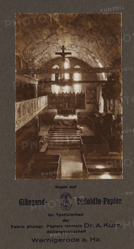 Interior of St. John's Church in Wernigerode, photograph, page, photographer: Hermann Günther, Wernigerode, 1923 - 1928, Hermann Günther, photographer, Saxony-Anhalt, paper, baryta paper, daylight collodion silver print, height 135 mm x width 88 mm, interior of church, St. John's Church, A sepia-toned interior photograph shows a vaulted stone ceiling with visible masonry and arched windows at the far end, through which bright light filters into the space. A large crucifix hangs from a horizontal beam near the ceiling, silhouetted against the light. Below, an altar covered with a draped cloth stands in front of an ornate altarpiece composed of framed panels and decorative pinnacles. Wooden pews and benches are arranged in rows leading toward the altar, and carved wooden galleries or paneling run along the left wall. Decorative elements, framed pictures, and sculptural details appear along the side walls, and a pulpit or carved wooden structure is visible on the right. The photograph is ...