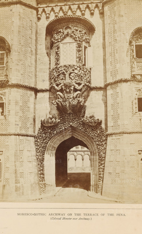 Entrance gate on the terrace of the Palácio da Pena in Sintra, Moresco-gothic archway on the terrace of the Pena (Colossal Monster over Archway), photograph, page, Sintra, c. 1865 - in or before 1870, photographer, paper, albumen print, height 197 mm x width 142 mm, Photo on third unnumbered page after page 20., gate, entrance, palace, Palácio da Pena, A sepia-toned photograph of a decorated stone archway set into an ornate façade, with a pointed arch passageway leading inward to a view of arched openings beyond; above the arch is a sculptural relief of a grotesque human-like face with muscular arms and bent legs emerging from a shell-like base, surrounded by carved foliage and wave-like ornamental forms; flanking the central bay are textured stone towers and patterned wall surfaces with framed windows and decorative stonework, and the whole composition is richly detailed with relief carvings and ornamental borders.