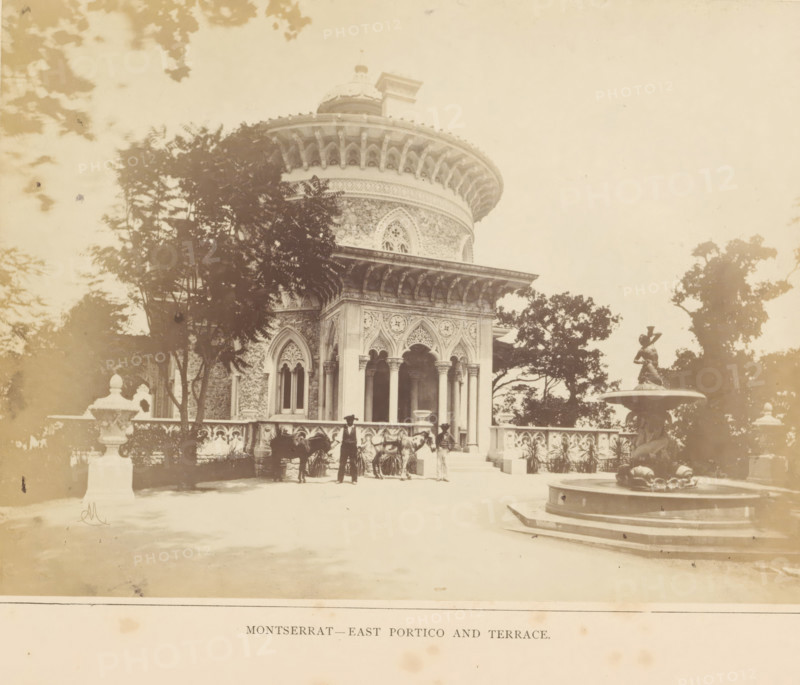 View of the east portico of the Palácio de Monserrate near Sintra, Montserrat - East Portico and Terrace, photograph, page, Sintra, c. 1865 - in or before 1870, photographer, paper, albumen print, height 149 mm x width 200 mm, palace, terrace, gate, entrance, Palácio de Monserrate, A pale, sepia-toned photograph shows an ornate round building with a domed roof and decorative arches, partially obscured by trees in the foreground; the building features carved stone columns, arched windows with tracery, and an overhanging cornice with regularly spaced brackets. In front of the building a balustraded terrace runs along the facade, with a group of people standing near the entrance and a horse present beside them. On the right a multi-tiered fountain with a sculpted figure stands on a raised circular base, and large trees frame the scene on both sides. The overall image has a faded, vintage appearance with soft shadows and scattered foliage.