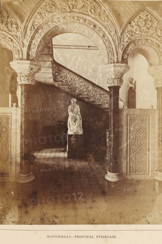 Stairwell in the Palácio de Monserrate near Sintra, Montserrat - Principal Staircase, photograph, page, Sintra, c. 1865 - in or before 1870, photographer, paper, albumen print, albumendruk, height 199 mm x width 146 mm, piece of sculpture, reproduction of a piece of sculpture, staircase, Palácio de Monserrate, A sepia-toned interior view showing a carved stone staircase with an ornate balustrade rising between sculpted archways, fluted columns with decorated capitals, patterned floor tiles, a seated marble statue on a pedestal positioned beneath the staircase, a standing bust displayed in a niche, and intricately patterned wooden or stone screens flanking the space.