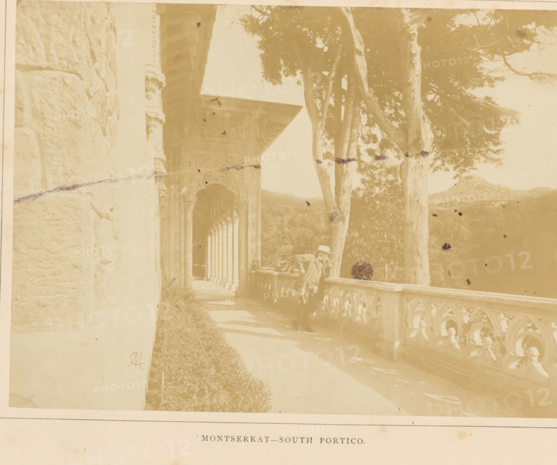 South portico of the Palácio de Monserrate near Sintra, Montserrat - South Portico, photograph, page, monogram: 'M', Sintra, c. 1865 - in or before 1870, photographer, paper, albumen print, albumendruk, height 146 mm x width 199 mm, colonnade, palace, gate, entrance, Palácio de Monserrate, A sepia-toned photograph showing an ornate stone portico with a row of carved columns and arches receding into the background beside a balustraded terrace, a man wearing a hat leaning on the terrace railing, tall trees with textured trunks rising beside the terrace, a stone wall at the left edge, decorative architectural details around the archway, shadows cast on the walkway, and distant hills visible beyond the trees.