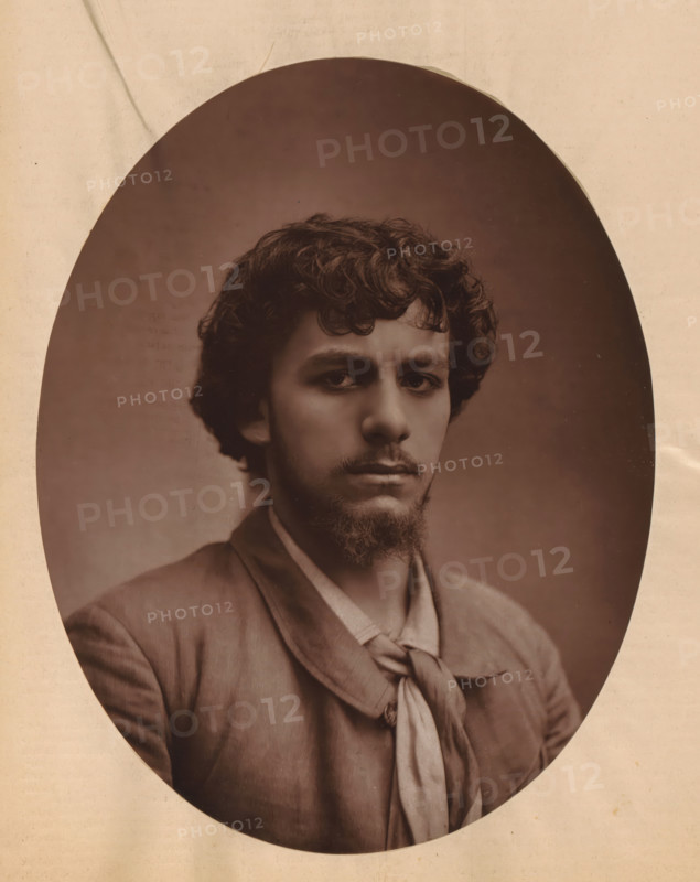 Portrait of Albert Lambert, page, photomechanical print, photographer: Félix Nadar, cliché maker: Lemercier, c. 1880 - c. 1885, paper, Woodburytype, Woodburytypepie, height 243 mm x width 189 mm, Person, Albert Lambert, historical persons, portrait of actor, actress, A sepia oval portrait shows a young man with curly dark hair and a short beard and mustache, his face turned slightly toward the camera with a neutral expression, wearing a jacket over a shirt and a loosely knotted scarf around his neck, set against a plain darker background and framed by a lighter paper border with subtle creases and spots.