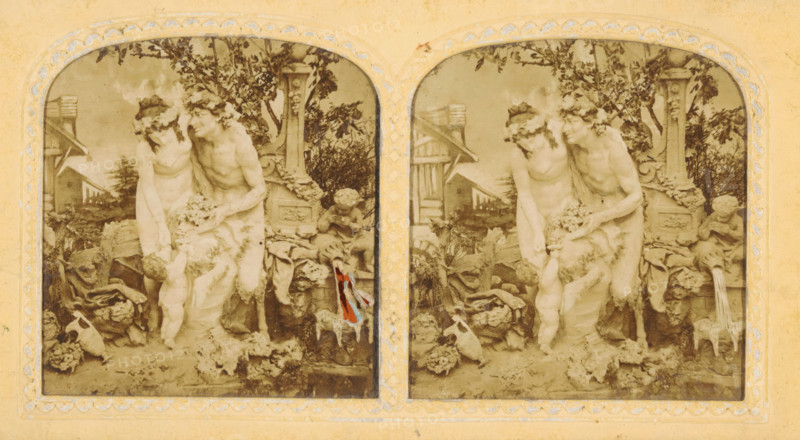 Mythological figures, presumably made of wax, for a backdrop, tissue stereograph, Europe, c. 1850 - c. 1880, photographer, paper, pasteboard, transparent paper, albumen print, hand coloring, pasted onto hand-colored transparent paper, between die-cut and embossed cardboard, height 85 mm x width 170 mm, cupids: 'amores', 'amoretti', 'putti', satyr(s) (in general), A sepia-toned stereoscopic image shows two arched panels side by side, each depicting a sculptural scene of a seated male figure wearing a garland and a seated female figure leaning toward him, both draped with cloth and surrounded by foliage and garden elements. A small childlike figure stands at their feet reaching upward toward the draped cloth, while another childlike figure sits nearby holding something close to its face. The background contains leafy branches, a decorative stone pillar with carved details, and a wooden structure partially visible behind the figures. Various objects and textured elements lie around the ba...