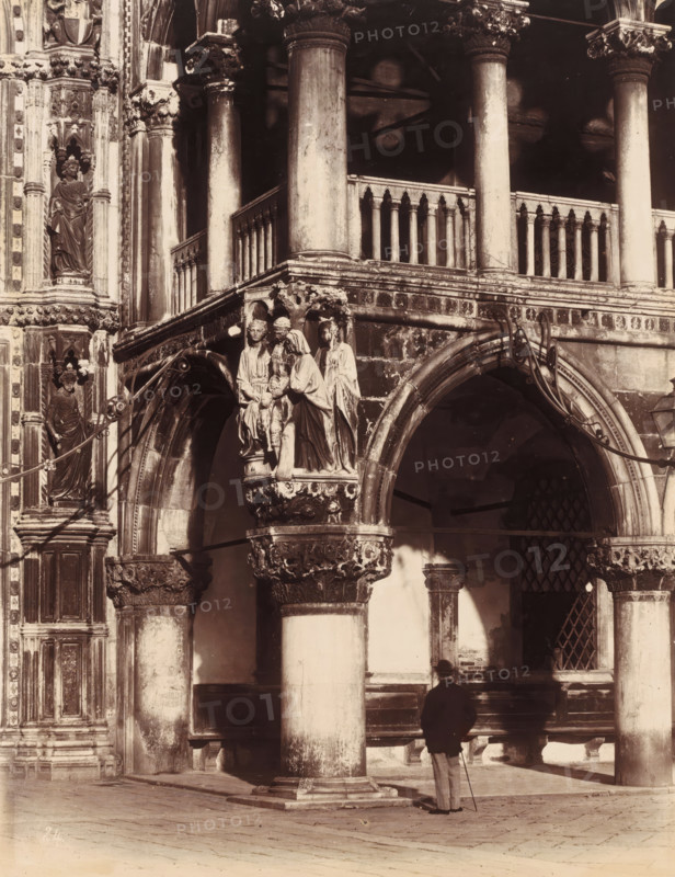 Palazzo Ducale, photograph, c. 1850 - c. 1870, photographer, albumen print, height 583 mm x width 446 mm, height 346 mm x width 267 mm, Venice, Doge's Palace, A sepia-toned photograph of an ornate stone façade featuring tall columns, arched openings, carved capitals, and balustrades, with sculpted figures in relief mounted on a projecting bracket above an arch; a man wearing a dark jacket and light trousers stands with a cane at the base of a central column, casting a shadow on the wall behind him; decorative vertical panels with recessed niches and statues flank the left edge of the image; wrought metal brackets curve above the arches and a barred window is visible through one arch; the ground is paved and shows shadows from the architectural elements.