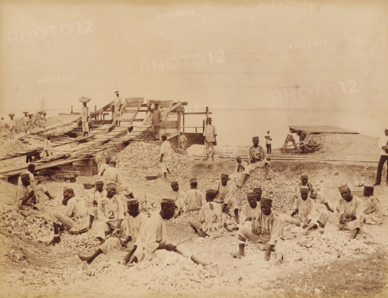United States, prisoners at Rock Fort Kingston (former title), Prisoners at Rockfort, Kingston, Jamaica, photograph, c. 1860 - c. 1890, photographer, albumen print, height 301 mm x width 396 mm, height 261 mm x width 338 mm, Rockfort, A wide sepia photograph shows a shoreline worksite with many men wearing light-colored loose clothing and caps seated on and standing among large piles of small rocks and gravel; several men hold hand tools while others sit on the gravel piles or rest on wooden planks. In the midground there are wooden ramps and sluice-like structures with slatted screens and chutes leading down to the piles, and a few men stand on and beside these structures, one carrying a rectangular tray on his head. Narrow rails and wooden platforms run across the site toward the water, and small wooden sheds and workbenches are visible near the right edge. The calm water fills the background horizon, and the sky above is pale and mostly empty. The image has an overall aged, grainy, ...