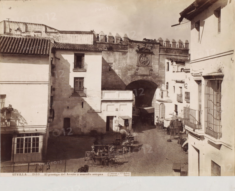 Sevilla-El postigo del aceite y muralla antigua, photograph, photographer: Juan Laurent, c. 1857 - c. 1880, albumen print, height 253 mm x width 337 mm, Andalusia, Seville, A sunlit street scene framed by whitewashed buildings with tiled roofs and iron balconies, showing a cobbled roadway leading toward a large arched gateway set into an old stone wall topped with crenellations, the gateway flanked by a decorative circular relief panel and sculpted details above it, market stalls and wooden tables displaying goods arranged along the street beneath awnings and cloth coverings, shadows cast across façades and the pavement, narrow windows and doorways punctuating the building faces, and various small architectural elements like cornices, drainpipes, and a barred ground-floor opening.