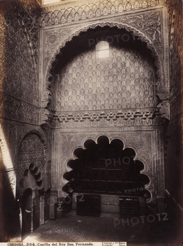 Cordoba-Capilla del Rey Don Fernando, photograph, photographer: Juan Laurent, c. 1857 - c. 1880, albumen print, height 334 mm x width 250 mm, Andalusia, Córdoba, An interior view showing a high decorative wall with an arched alcove and a small window near the top, surrounded by dense carved geometric and floral patterns, scalloped arch shapes framing a dark recessed area, ornate carved corbels and mouldings at the arch corners, tiled lower walls with patterned ceramic designs, repeating arabesque motifs covering the surfaces, shadowed recesses and niches, a patterned floor partially visible at the bottom, and strong contrasts of light and shadow across the carved surfaces.