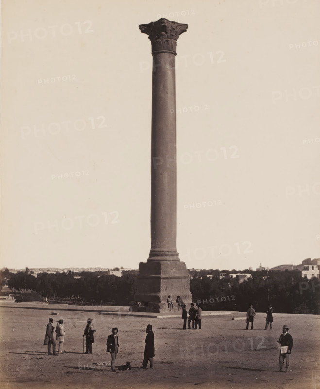 Pompey's Pillar, Alexandria, Pompey's Pillar at the Serapeum in Alexandria, photograph, c. 1870, photographer, North Africa, pasteboard, paper, albumen print, height 288 mm x width 355 mm, column, pillar, architecture, Serapeum, A tall single stone column rises from a square stepped base occupying the center of the scene, with a carved capital at its top. Trees and low buildings form a dark horizontal band behind the column. Several groups of people are scattered in the foreground around the base, wearing period clothing including long coats, hats, and some with walking sticks; a few individuals stand alone while others converse in small clusters. The ground in front of the column is bare and flat with faint shadows cast by the people. A small animal lies on the ground near one standing figure. The sky above the column is pale and empty.