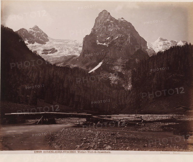 View of the Rosenhorn, Switzerland, Rosenlaigletscher. Wetter-Well und Rosenhorn, photograph, inscription: '1665', inscription: '150=', number: '12859', number: '140', photographer: Giorgio Sommer, Berner Alpen, 1857 - 1914, Alpen, pasteboard, paper, albumen print, albumendruk, height 333 mm x width 435 mm, brook, dale, valley, mountain-top, peak, Alps, Bernese Alps, Rosenhorn, A wide sepia-toned landscape photograph showing a steep rocky mountain with patches of snow rising behind a dense dark forest of tall conifer trees, flanked by additional snow-capped peaks and a glacier visible on one side, with a shallow river or stream in the foreground flowing past a rocky, pebble-strewn bank and a simple log bridge spanning the water.