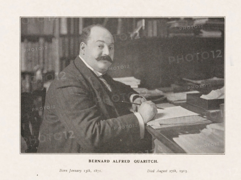 Bernard Alfred Quaritch, Portrait of bookseller Bernard Alfred Quaritch behind his desk, print, photograph, after 1913-08-27, printmaker, photographer, paper, printing, height 225 mm x width 287 mm, Person, book-shop, bookseller, A sepia-toned photograph of a seated man with a rounded face and a prominent mustache, wearing a dark suit and a white shirt collar, shown from the waist up at a desk covered with stacks of papers and folders; his right hand holds a pen above an open sheet while his left rests on the desk, and behind him are blurred bookshelves and indistinct objects on the surface behind the desk.