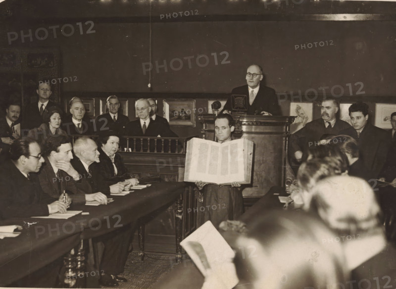 Auction of the Gutenberg Bible by Charles William Dyson Perrins at Sotheby's in London, photograph, Interior of the auction room at Sotheby's London during the auction of Charles William Dyson Perrins in 1947. In front of the auctioneer stands a holder holding the open Gutenberg Bible in his hands., label: 'Rarest edition of the scriptures bought for £ 22, 000, ', stamp: 'Copyright, Keystone Press Agency Ltd, annotation, photographer: Keystone Press Agency, 1947, Keystone Press Agency, photographer, paper, gelatin silver print, gelatin silver print on photographic paper, height 152 mm x width 206 mm, The Bible was sold for £22, 000 to Maggs Bros. The bids were placed by Ernest Maggs, the man with the goatee behind the table on the left., Person, Ernest Maggs, art sale, auction, Bible, A sepia-toned photograph of a room filled with seated and standing people around a long table, with a man standing behind a raised wooden lectern near the back of the room and another person standing in f...