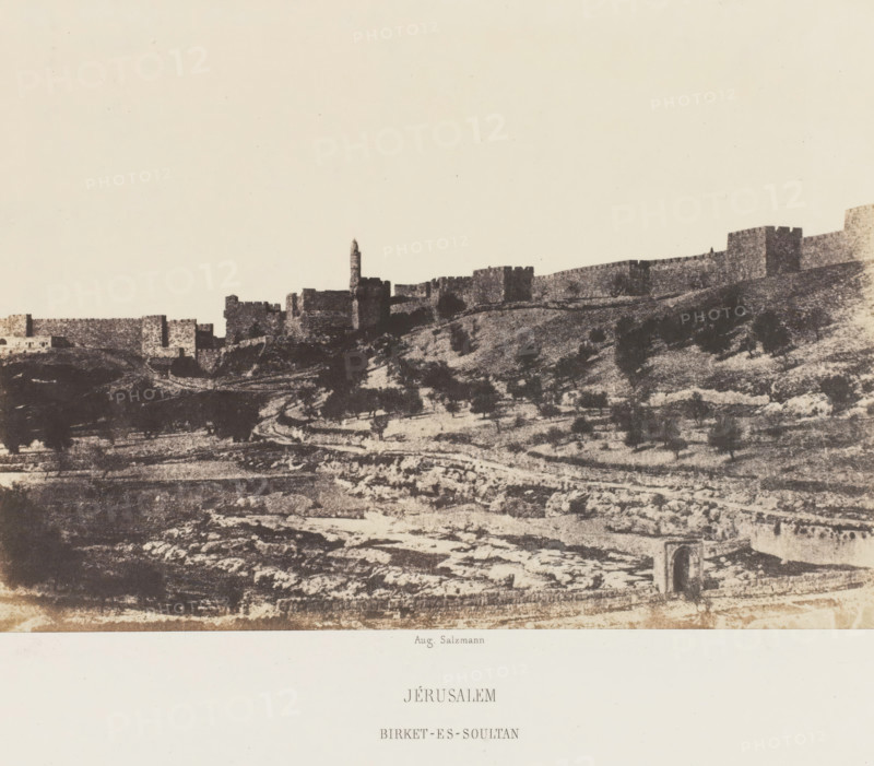 Basin of Birket-es-Soultan outside the Old City Wall of Jerusalem, Jérusalem. Birket-es-Soultan, photograph, page, photographer: Auguste Salzmann, Jerusalem, printer (person): Louis-Désiré Blanquart-Evrard, Lille, publisher: Gide et J. Baudry, Paris, 1854 - 1856, printer (person), pasteboard, paper, salted paper print, height 155 mm x width 218 mm, Photo on unnumbered page., city-view in general, 'veduta', pond, pool, Birket-es-Soultan, A wide sepia-toned landscape showing a stone city wall and fortress along a ridge with a tall narrow tower rising above the ramparts, the foreground featuring terraced, rocky hillside with scattered dark shrubs and a winding path, and an arched stone opening visible near the lower edge.