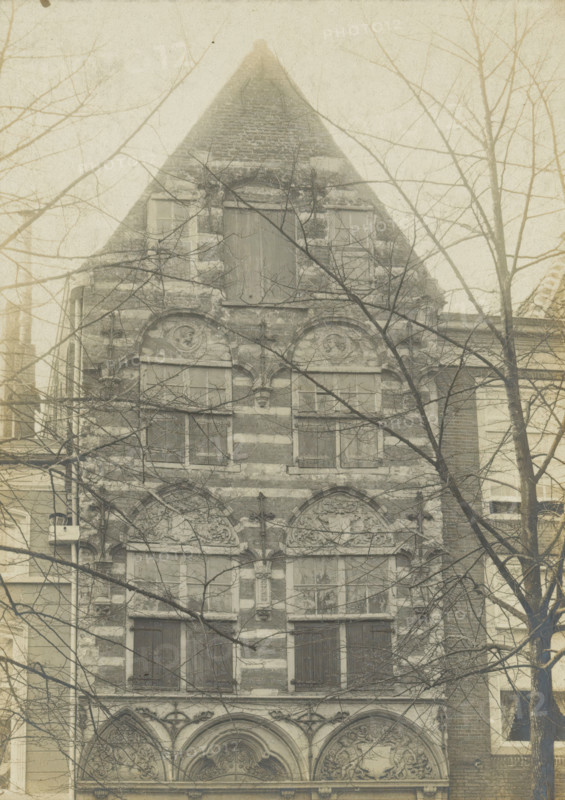 Facade of a house on the Koornmarkt in Delft, Koornmarkt WZ No. 81. Early Renaissance facade, photograph, number: '27', inscription: 'Delft. 1st half of the 16th century.', inscription: 'The right of reproduction of these photos is reserved by the Publisher.', Delft, c. 1900 - c. 1904, photographer, paper, pasteboard, gelatin silver print, height 167 mm x width 120 mm, urban housing, ornament derived from architectural details, Koornmarkt, A tall narrow building facade with a steep triangular roof, masonry bands visible across the exterior, multiple rectangular windows with some shutters closed, ornate arched decorative reliefs above lower windows, bare tree branches partially obscuring the view, adjacent building walls visible at the sides, and a muted, aged photographic tone.