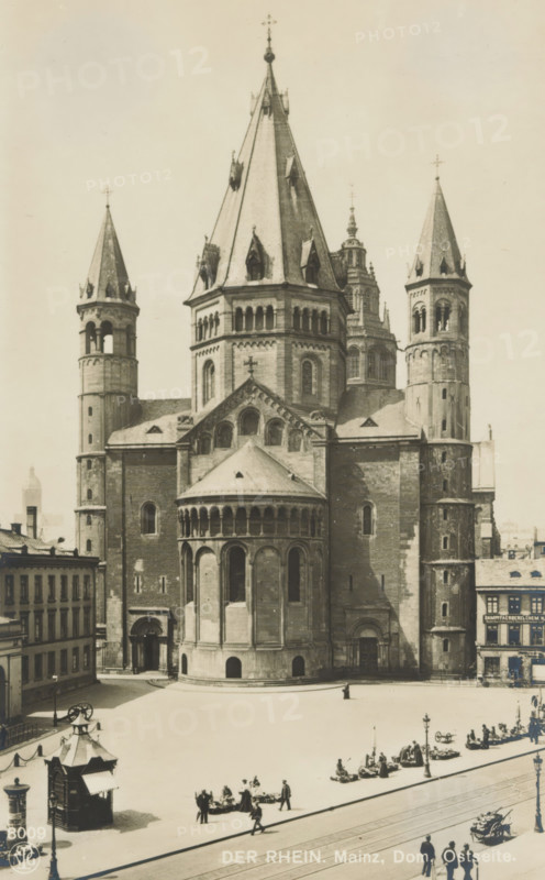 View of Mainz Cathedral, Der Rhein. Mainz, Dom, Ostseite, Bilder aus der Kunst aller Zeiten-Herausgeber: F. Grukendorf Mappe 5 Deutscher Kirchbau im Mittelalter, photograph, number: '3587', number: '8009', photographer: Neue Photographische Gesellschaft, Mainz, c. 1900 - in or before 1911, Rhineland-Palatinate, paper, pasteboard, gelatin silver print, developing gelatin silver print, height 138 mm x width 86 mm, church (exterior), parts of church exterior and annexes: tower, Mainz Cathedral, A large stone cathedral with multiple tall conical and pyramidal spires dominates the scene, featuring round towers with arched openings and a central apse with a semi-circular colonnaded exterior; the façade shows rows of arched windows and decorative stonework. In front of the building is a broad open square with small groups of people standing or sitting, streetlamps, and a few horse-drawn carriages; a small kiosk structure with a pitched roof is visible at the left foreground. Adjacent building...