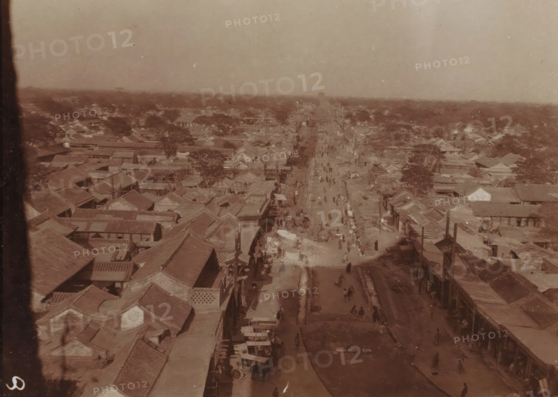 Bird's-eye view of a long shopping street in a vast city, China, photograph, inscription: 'Douairière Hoeufft-van de Spie etc. wedding announcement', photographer: attributed to Jan Adriani, 1907, Jan Adriani, photographer, Attributed to, paper, daylight collodion silver print, daylight collodion silver print on photo paper, height 80 mm x width 105 mm, city-view in general, 'veduta', street, A sepia-toned aerial view of a wide street running through a dense area of low buildings with tiled roofs, the street lined with small structures and stalls and dotted with many tiny figures walking or standing, trees scattered among the rooftops, a distant hazy horizon, and a vertical dark strip along the left edge of the image.