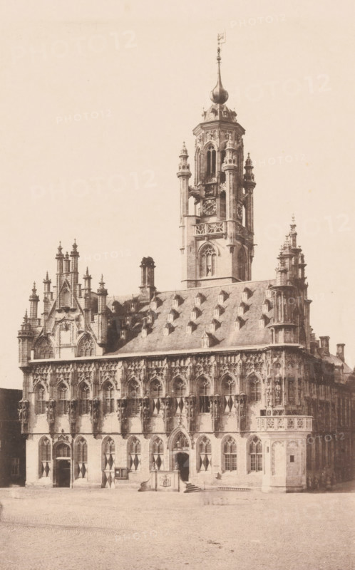 View of the Town Hall of Middelburg, photomechanical print, c. 1850 - c. 1900, paper, collotype, photogravure, height 261 mm x width 188 mm, town hall, Middelburg, Stadhuis (Middelburg), A sepia-toned photograph of an ornate Gothic-style building with a large central tower rising above a steeply pitched roof dotted with small dormer windows, the tower featuring arched openings, decorative columns, a round clock face, and a bulbous spire topped by a finial; the facade below displays tall arched windows with tracery, sculpted figures in niches, pinnacles, and decorative stonework, while the ground level shows arched doorways with patterned shutters and a small raised platform or entrance feature with steps in front; the foreground is a cobbled open area.
