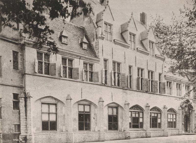 View of the Abbey of Middelburg from the inner courtyard, Former residence of the abbot, Ancien logement de l'abbé, Views of the Abbey in Middelburg, Abbey in Middelburg, Abbaye de Middelbourg, photomechanical print, cliché maker: Straaten, Gebroeders van, Middelburg, in or after 1900, Straaten, photographer, cliché maker, paper, collotype, photogravure, height 150 mm x width 202 mm, abbey, monastery, convent, Roman Catholic church, Abbey of Middelburg, A stone building facade with arched ground-level windows and rectangular upper windows, some of which have open wooden shutters; a steep tiled roof with small dormer windows and larger gabled dormers above; decorative molding and stone trim around windows and along the roofline; leafy tree branches partially framing the top corners of the image; a paved area in front of the building.