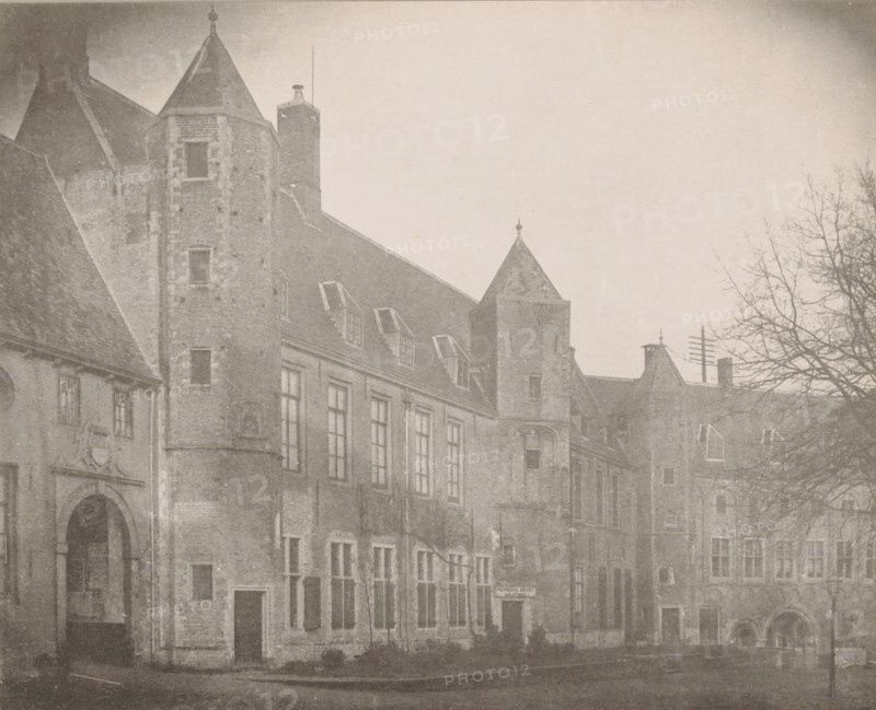 View of the State Archives buildings in the Abbey in Middelburg, The State Archives buildings on the north side of the large courtyard in 1888, The buildings of the State Archives, Views of the Abbey in Middelburg, Abbey in Middelburg, Abbaye de Middelbourg, photomechanical print, cliché maker: Straaten, Gebroeders van, Middelburg, in or after 1900, photographer, paper, collotype, photogravure, height 150 mm x width 186 mm, courtyard, abbey, monastery, convent, Roman Catholic church, archive, Abbey of Middelburg, A sepia-toned photograph of an old stone building with steep pitched roofs and multiple towers, showing rows of tall windows along the facade and several dormer windows set into the roof; a large arched doorway is visible at one end and smaller doorways and window openings punctuate the ground level; a bare-branched tree stands on the right side of the scene, its limbs extending into the sky above the roofline; portions of the street and pavement run in front of the building.