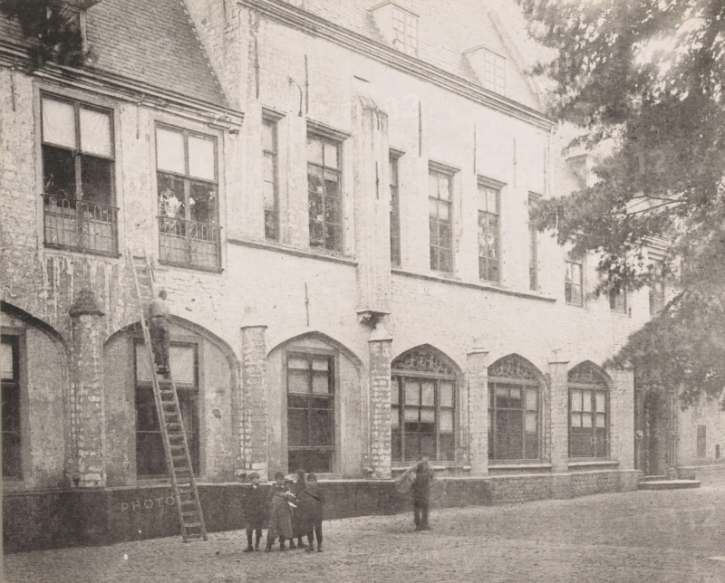View of the former residence of the abbot in the inner courtyard of the Abbey in Middelburg, 1894, Former residence of the abbot, later of the bishop, Ancien logement de l'abbé, Views of the Abbey in Middelburg, Abbey in Middelburg, Abbaye de Middelbourg, photomechanical print, cliché maker: Straaten, Gebroeders van, Middelburg, in or after 1900, Straaten, photographer, cliché maker, paper, collotype, photogravure, height 149 mm x width 185 mm, courtyard, abbey, monastery, convent, Roman Catholic church, Abbey of Middelburg, A faded façade of a multi-storey building with tall rectangular windows set above a row of arched openings, rough stone walls and visible mortar, and decorative ironwork on some window ledges; a long ladder leaning against the wall reaching toward an upper window where a person stands partway up; a small group of children standing together in the courtyard in front of the building and another person standing nearby facing toward them; a tree with dense foliage part...