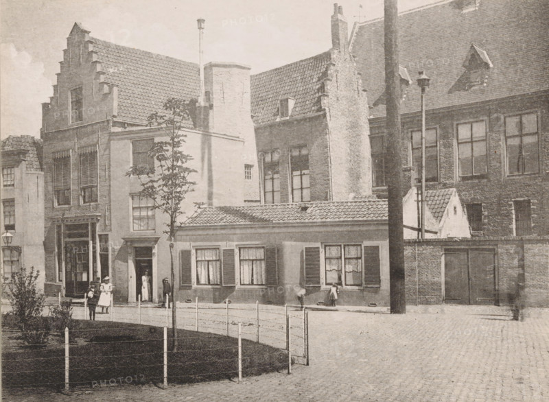 House on the Balance, purchased for the Provincial Water Authority, before the restoration, 1895, Maison sur la place de la Balance, View of the House on the Balance near the Abbey of Middelburg, Views of the Abbey in Middelburg, Abbey in Middelburg, Abbaye de Middelbourg, photomechanical print, cliché maker: Straaten, Gebroeders van, Middelburg, in or after 1900, photographer, paper, collotype, photogravure, height 150 mm x width 201 mm, abbey, monastery, convent, Roman Catholic church, Abbey of Middelburg, A row of brick and stucco buildings with tiled roofs and stepped gables lines a cobblestone street, several windows with shutters and lace curtains visible on the lower facades and tall multi-pane windows on the upper floors, a narrow doorway with a person standing inside and a few people—some wearing dresses and hats—standing and walking on the sidewalk, a small fenced grassy area with a young tree in front, a low building with shuttered windows and a tiled roof at street level, a...