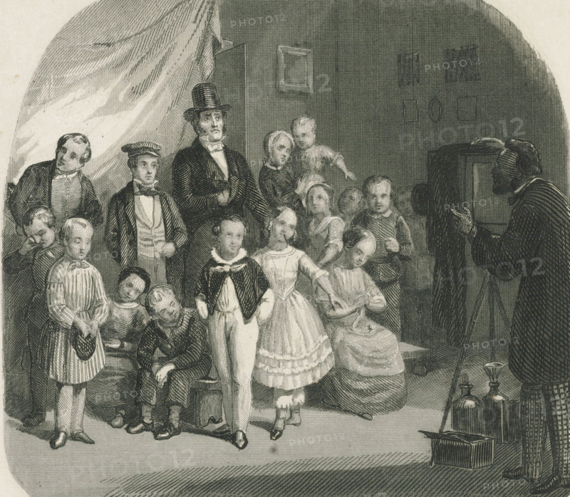 Family posing for a group photo, print, album page, Print is part of an album. Print is loose in the album next to the design drawing., printmaker: Johann Wilhelm Kaiser (I) (possibly), 1840 - 1900, paper, etching, height 126 mm x width 138 mm, family group, especially parents with their child(ren), taking photographs of model, sitter, camera, photography, A monochrome engraving showing a group of people gathered in an interior space, including men, women, and children in period clothing; a tall man wearing a top hat and formal coat stands near the center with a hand tucked to his chest, while a woman and a child stand close beside him and another woman sits holding fabric in her lap; several boys and girls in dresses and jackets occupy the foreground, some standing and some seated, with varied facial expressions and postures; on the right edge a bearded man in a dark robe faces the group and gestures with one hand, and near him on the floor are glass bottles and a small box with a lon...