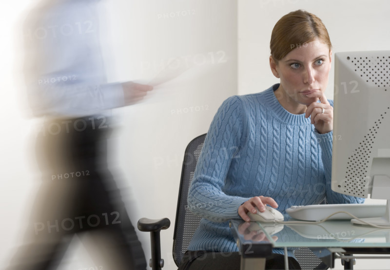 Woman at computer with person passing.