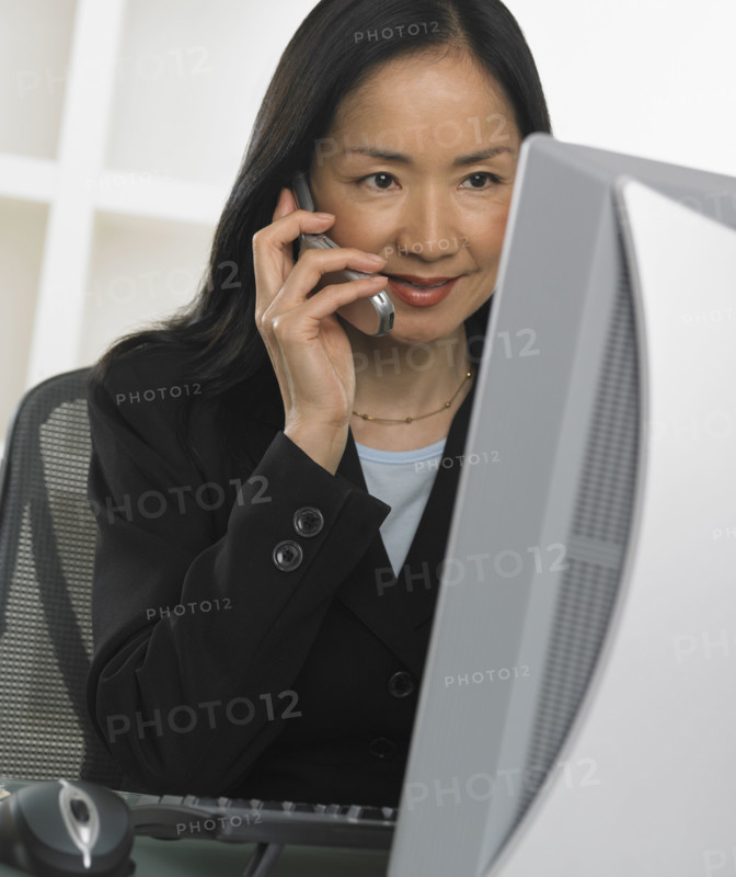 Asian businesswoman using cell phone at computer.