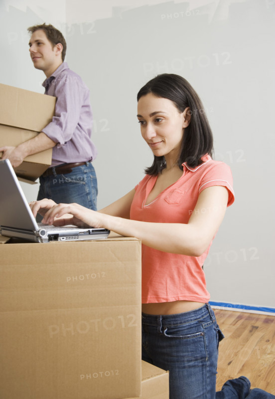 Woman using laptop while man carries box in new house. Date : 2006