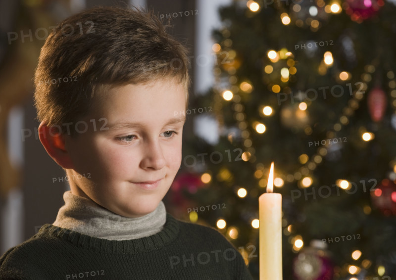 Boy looking at candle in front of Christmas tree.