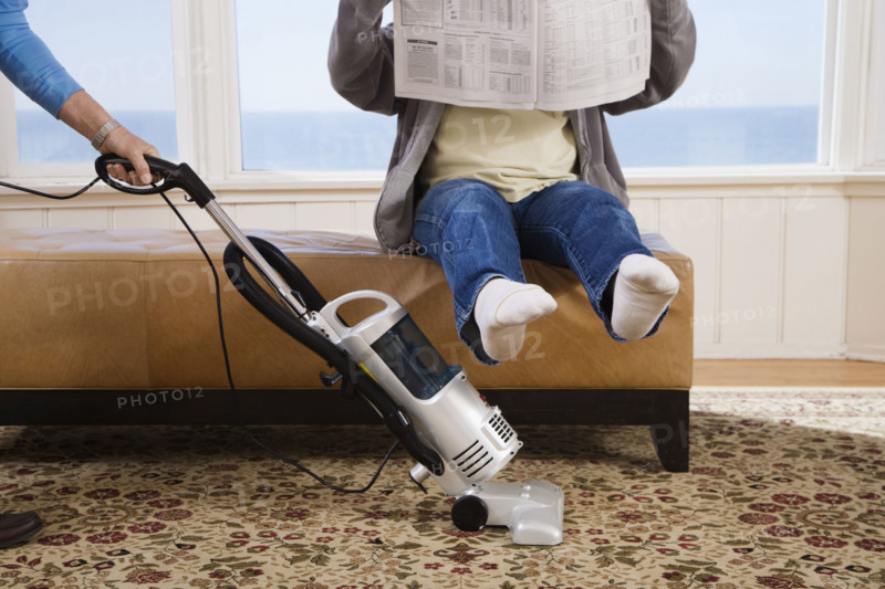 Woman vacuuming under man’s feet. Date : 2008