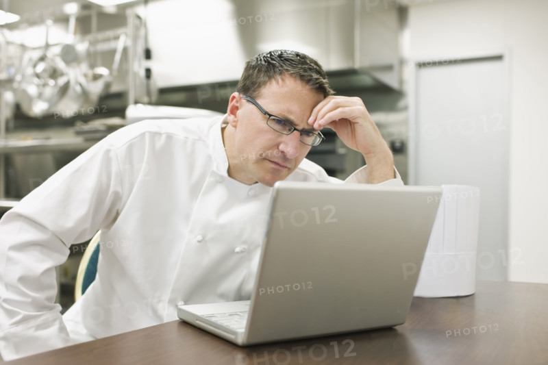 Frustrated chef looking at laptop in kitchen. Date : 2008