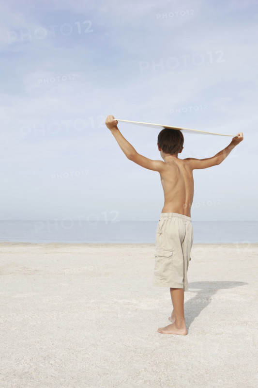 Boy standing on beach holding bodyboard overhead. Date: 2008