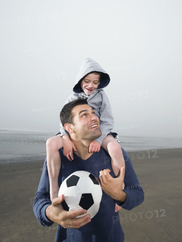 Father carrying son on shoulders and holding soccer ball on beach. Date : 2008