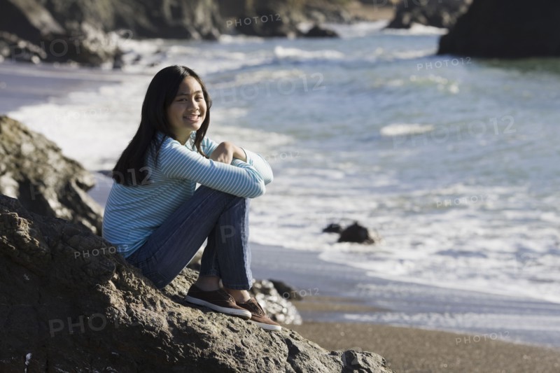 Teenage girl sitting on coastal rock. Photographe : PT Images