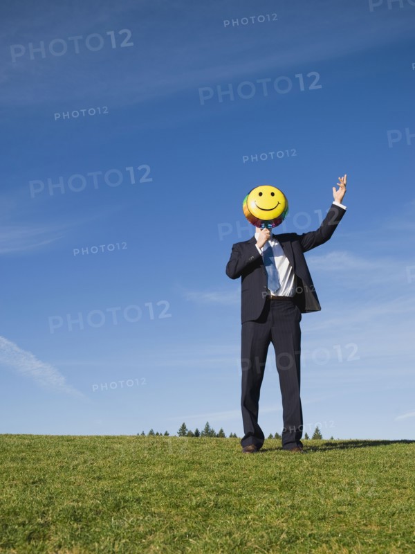 Businessman holding smiley face balloon over face.