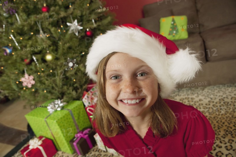 Girl (9) wearing Santa hat sitting in front of Christmas tree, portrait. Photographe : Sarah M. Golonka