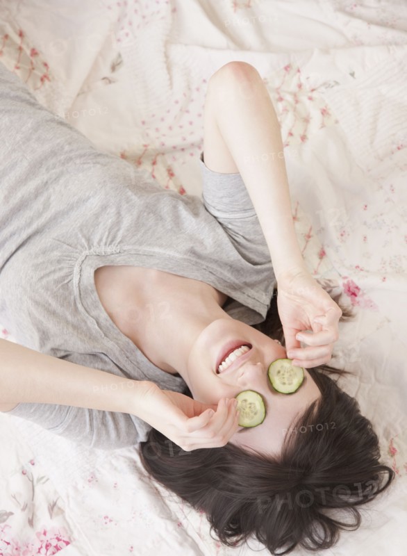 Young woman lying in bed with cucumber face mask. Photographe : Jamie Grill