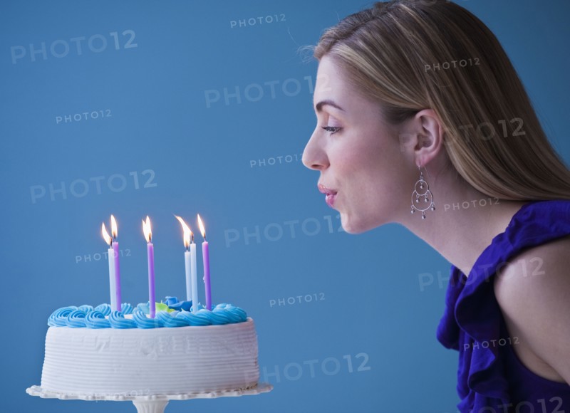 Young woman blowing candles on birthday cake, studio shot. Photographe : Daniel Grill