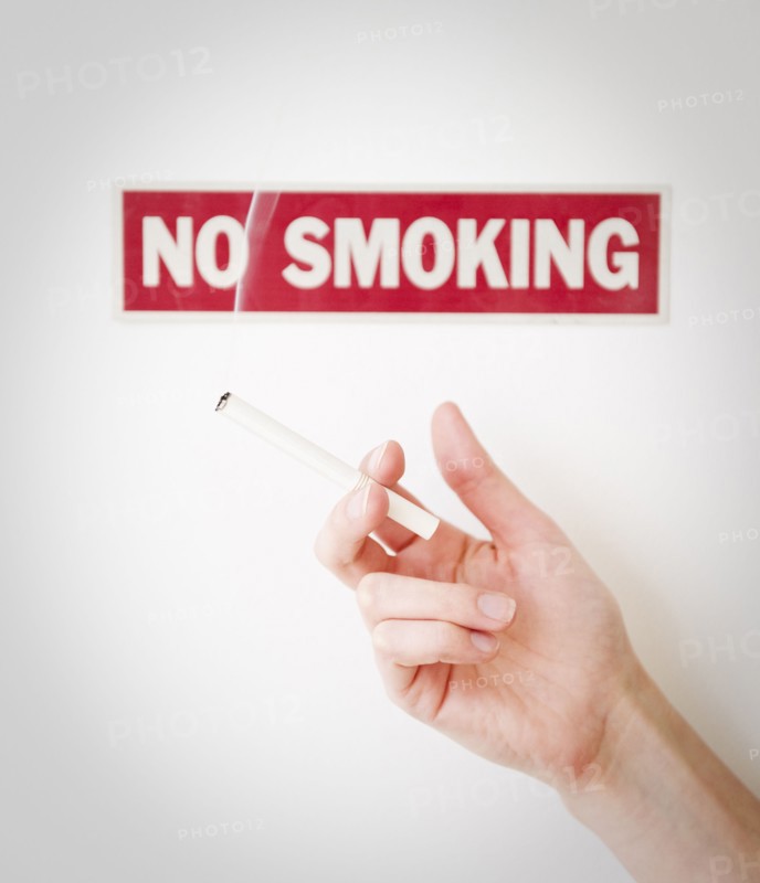 Hand holding cigarette in front of no smoking sign. Photographe : Jamie Grill