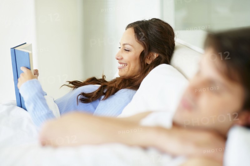 Woman reading book in bed beside sleeping husband. Photo. momentimages
