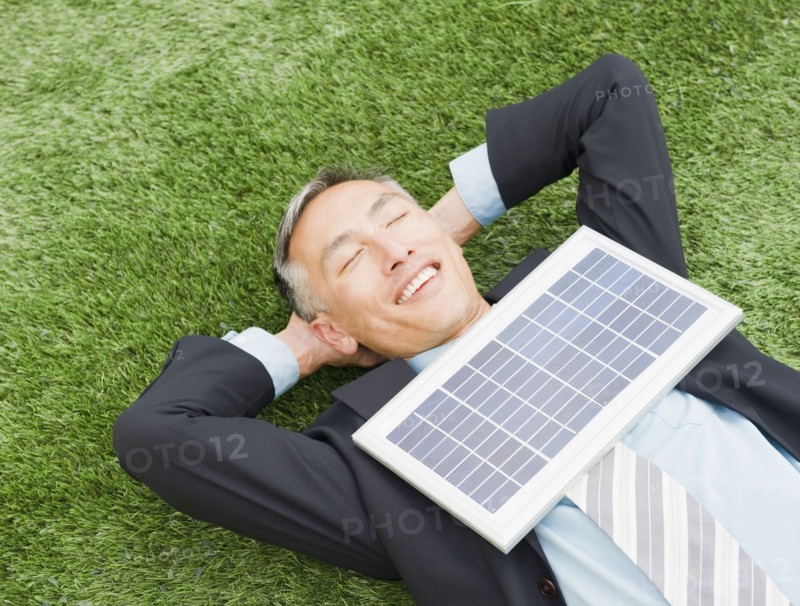 Happy businessman lying on grass with eyes closed and hands behind head and with solar panel on chest. Photo : Jamie Grill